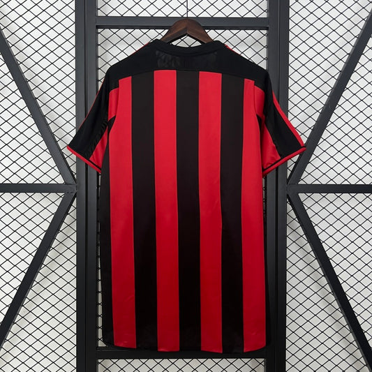Black and red Ac Milan 04/04 home football shirt on a hanger against a geometric patterned wall.
