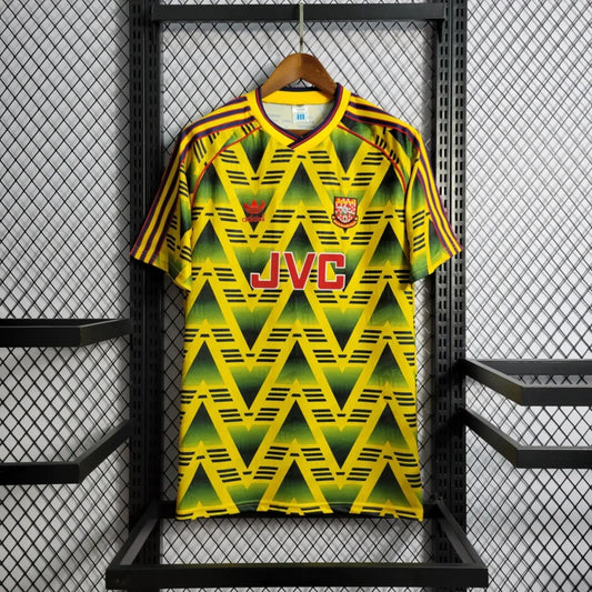 Yellow and green Arsenal 01-03 away shirt with 'JVC' branding on a hanger against a metallic background