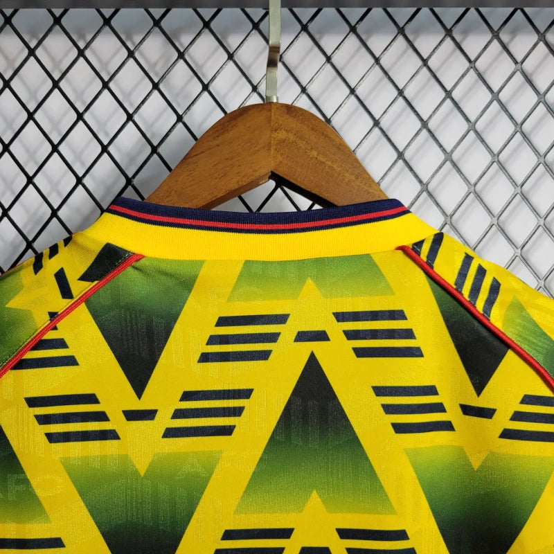 Yellow and green Arsenal 01-03 away shirt with black patterns on a wooden hanger against a chain-link fence background.