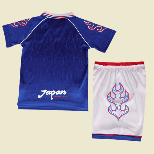 Blue Japan 1998 home kids kit with flame design and white shorts with red trim on a textured gray background
