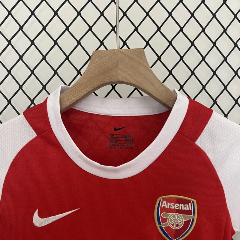 Red and white Arsenal 02-04 home kids kit with Arsenal logo on a hanger against a wire fence background