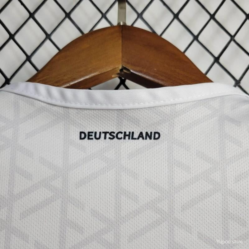 White Germany 2024 home baby onesie with 'DEUTSCHLAND' text on a wooden hanger against a chain-link fence background.