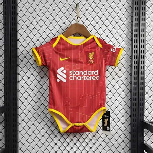 Red Liverpool 24/25 home baby onesie with yellow accents and sports logos on a hanger against a grid-patterned wall.