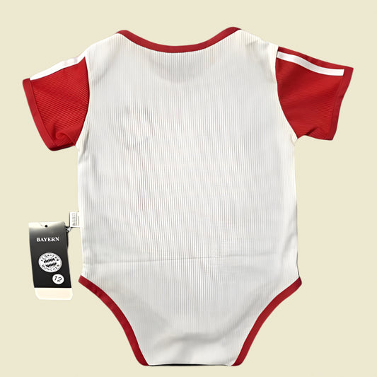 White Bayern Munich 23/24 home baby onesie with red sleeves and trim, featuring a Bayern Munich tag on a black background.