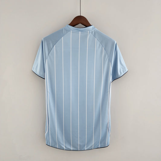 Light blue Man City 07/08 home shirt with vertical stripes on a hanger against a beige background