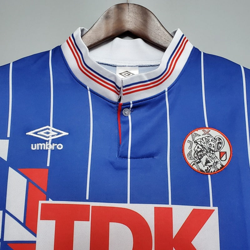 Blue Ajax 89/90 away shirt with red and white accents on a hanger, featuring Umbro logo and TDK branding.