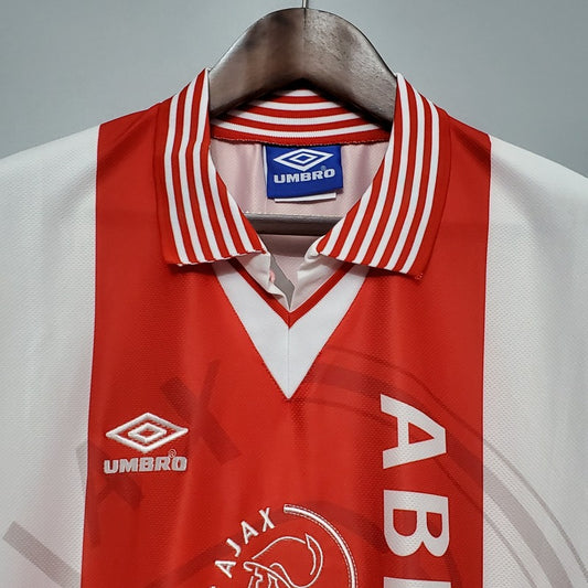 Red and white Ajax 95/96 home shirt with 'Umbro' and 'Ajax' logos on a hanger against a gray background.
