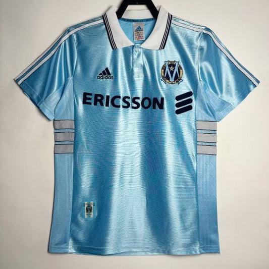 Light blue Marseille 98/99 away shirt with Adidas logo and Ericsson branding on a hanger against a light background