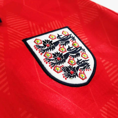 Close-up of a red England 1990 away shirt with a prominent emblem on a white background