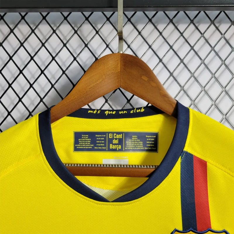 Yellow Barcelona 08/09 long sleeve away shirt with a red and blue stripe on a wooden hanger against a chain-link fence background.