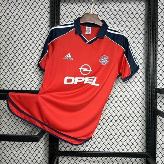 Red Bayern Munich 00/01 home shirt with navy accents, featuring Adidas and Opel logos, on a hanger against a wire mesh background.