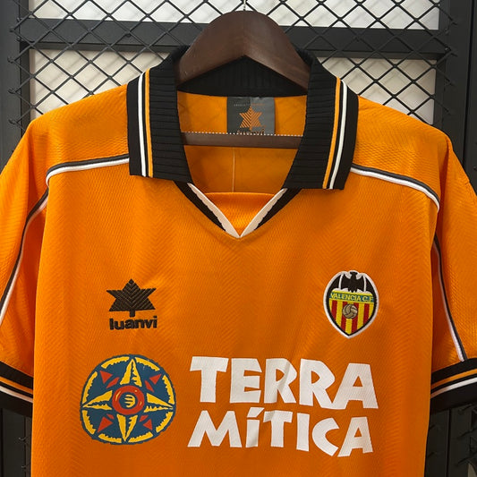 Orange Valencia 99/00 away shirt with branding and text on a hanger against a black metal fence background