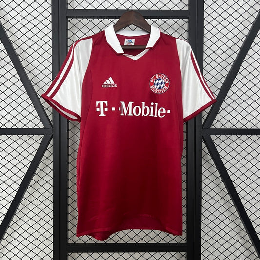 Red Bayern Munich 03/04 home shirt with white accents on a hanger against a geometric patterned background