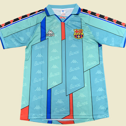 Light blue Barcelona 96/97 away shirt with red and black accents, featuring Barca logo, on a gray textured background.