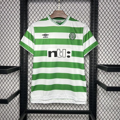 Green and white striped 99/00 Celtic home shirt with visible branding on a hanger against a black grid background.