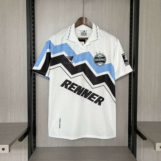 White 95/96 Gremio away shirt with blue and black zigzag pattern, featuring 'Renner' and 'Gremio' logos, hanging in a locker room.