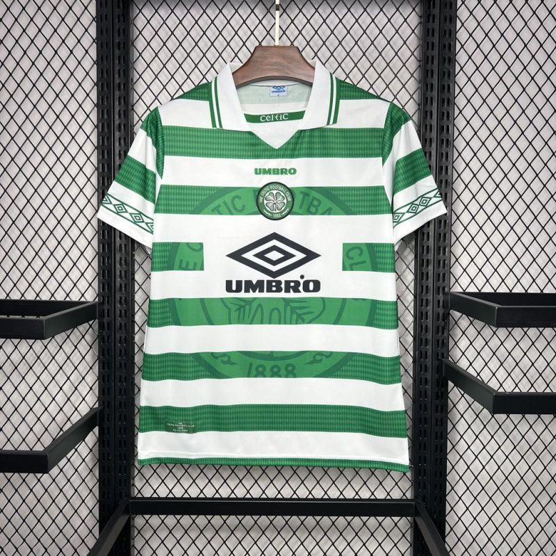 Green and white striped Celtic 98/99 home shirt with Umbro logo on a hanger against a black metal fence background.