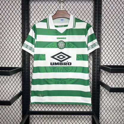 Green and white striped Celtic 98/99 home shirt with Umbro logo on a hanger against a black metal fence background.