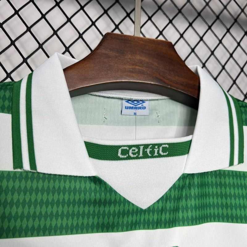 Green and white Celtic 98/99 home shirt with 'Celtic' branding on a hanger against a chain-link fence background.