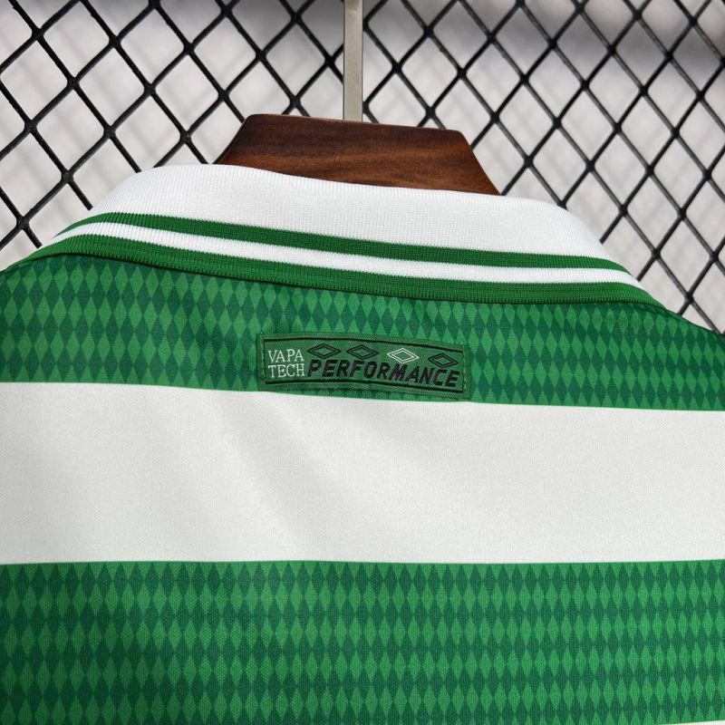 Green and white Celtic 98/99 home shirt with 'Vapa Tech Performance' label on a hanger against a chain-link fence background.