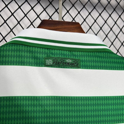 Green and white Celtic 98/99 home shirt with 'Vapa Tech Performance' label on a hanger against a chain-link fence background.