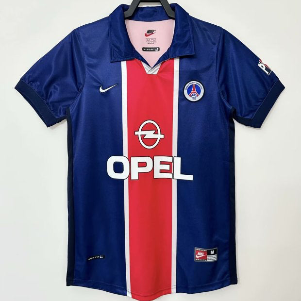 Blue psg 98/99 home shirt  with red and white stripe, featuring Opel logo, on a hanger against a white background.