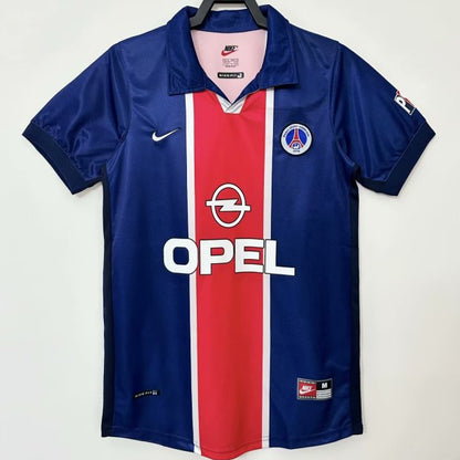 Blue psg 98/99 home shirt  with red and white stripe, featuring Opel logo, on a hanger against a white background.