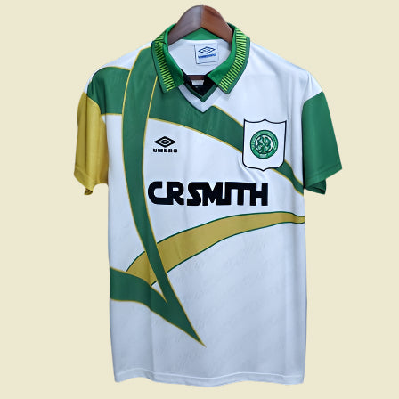 Green and white 93-95 Celtic away shirt with yellow accents on a hanger against a black background