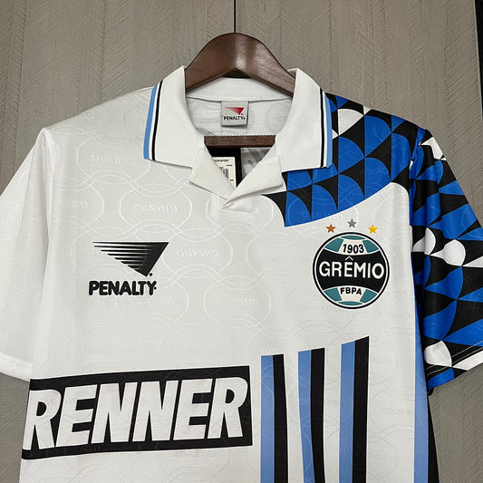 White Gremio 94/95 away shirt with blue and black accents, featuring logos on a hanger against a neutral background.