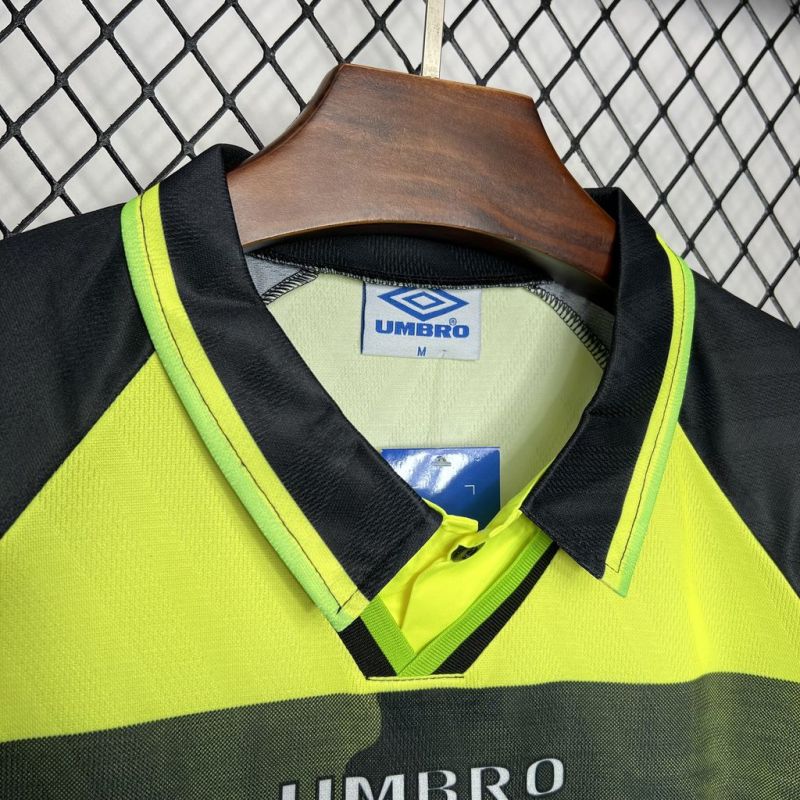 Yellow and black Celtic 96/97 away kit with Umbro logo on a hanger against a chain-link fence background.