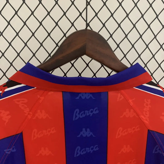 Red and blue Barcelona 96/97 long sleeve home shirt with 'Barça' branding on a hanger against a chain-link fence background.