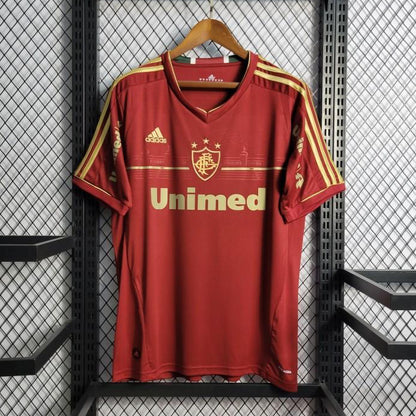 Red Fluminense 2012 third shirt  with gold accents on a hanger against a black metal cage background