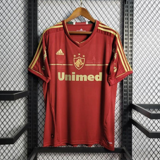 Red Fluminense 2012 third shirt  with gold accents on a hanger against a black metal cage background