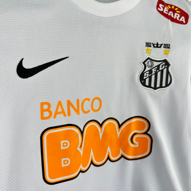 White Santos 2012 centenary home kids kit with Nike logo, S.C.G. emblem, and 'Banco BMG' branding.