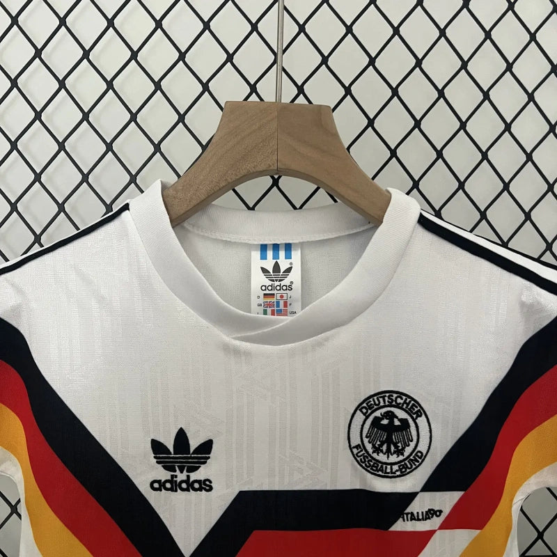 White Germany 90 World Cup home kids kit  with black, red, and yellow stripes on a hanger against a patterned wall.