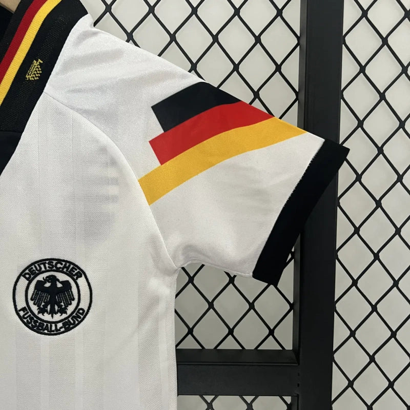 White Germany 1992 home kids kit with German flag design and logo on a black stand against a wire mesh background.
