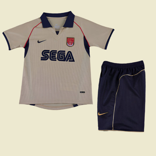 Beige Arsenal 01/02 away kids kit with navy accents and Sega logo, paired with navy shorts on a textured gray background.