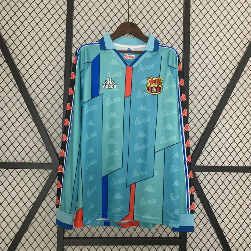 Teal Barcelona 96/97 long sleeve away shirt  with red and blue accents, featuring a logo, on a hanger against a wire mesh background.