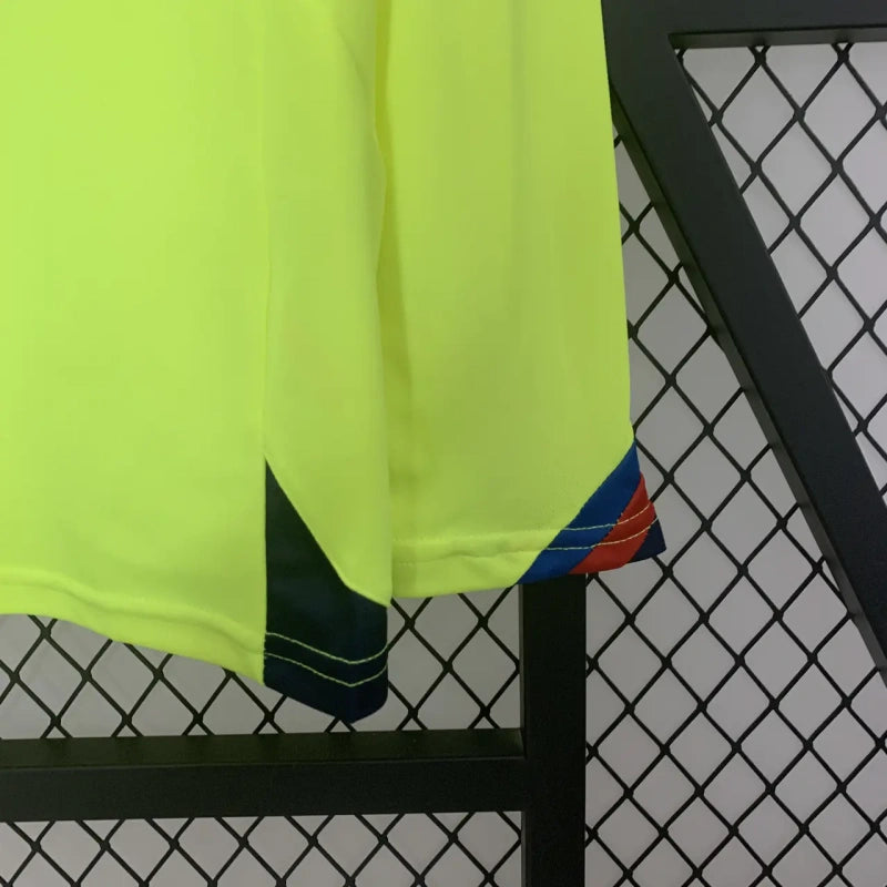Bright yellow Barcelona 05/06 long sleeve away shirt with black and blue accents hanging on a black metal rack against a chain-link fence.