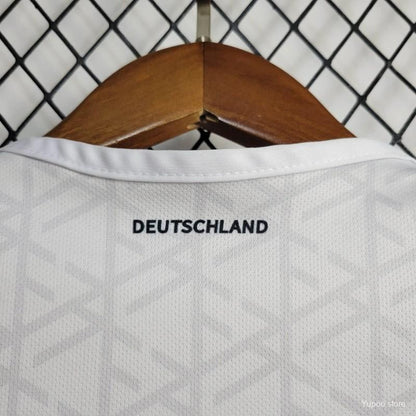 White Germany 2024 home baby onesie  with 'DEUTSCHLAND' text on a wooden hanger against a chain-link fence background.