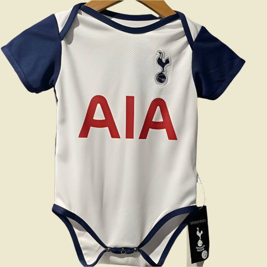 White Tottenham 24/25 home baby onesie with blue sleeves and AIA logo on a hanger against a gray wall.
