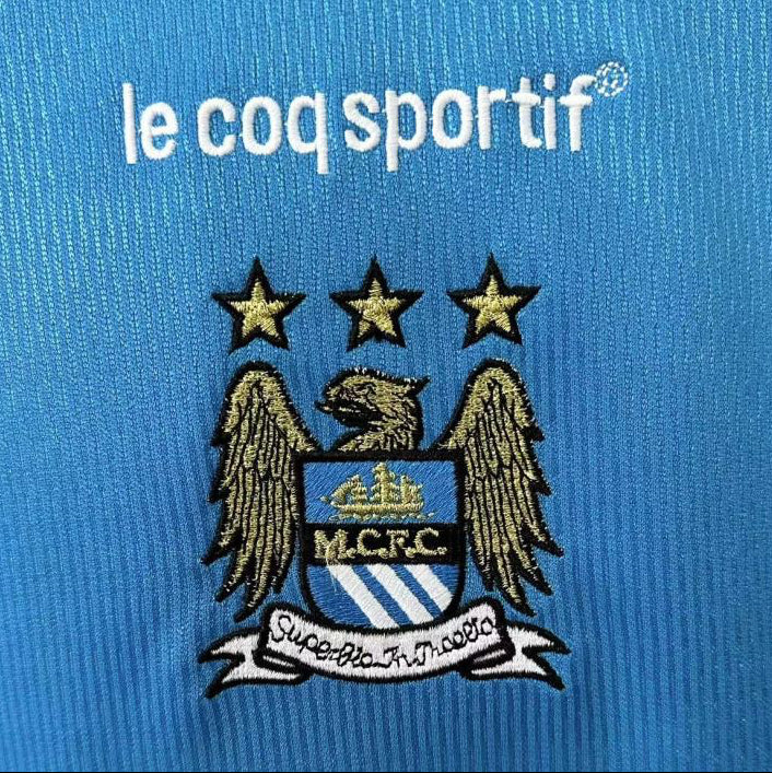 Manchester City logo with 'le coq sportif' on a blue background