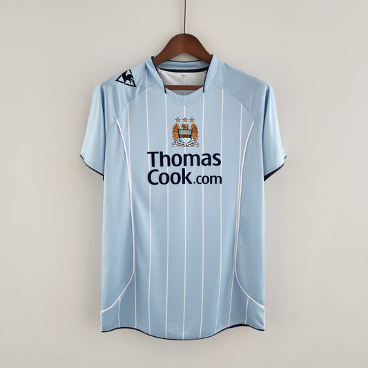 Light blue Man City 07/08 home shirt with 'Thomas Cook.com' text on a hanger against a light gray background
