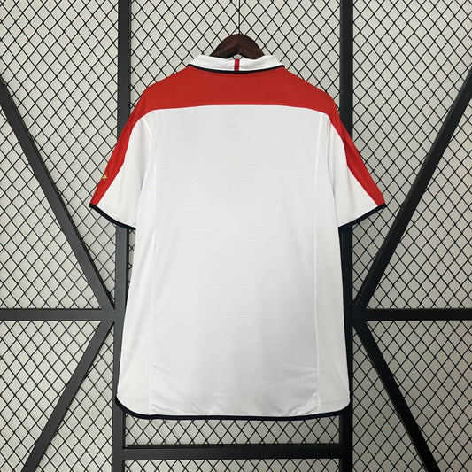 White England 2004 home shirt with red accents on a black metal stand against a wire mesh background